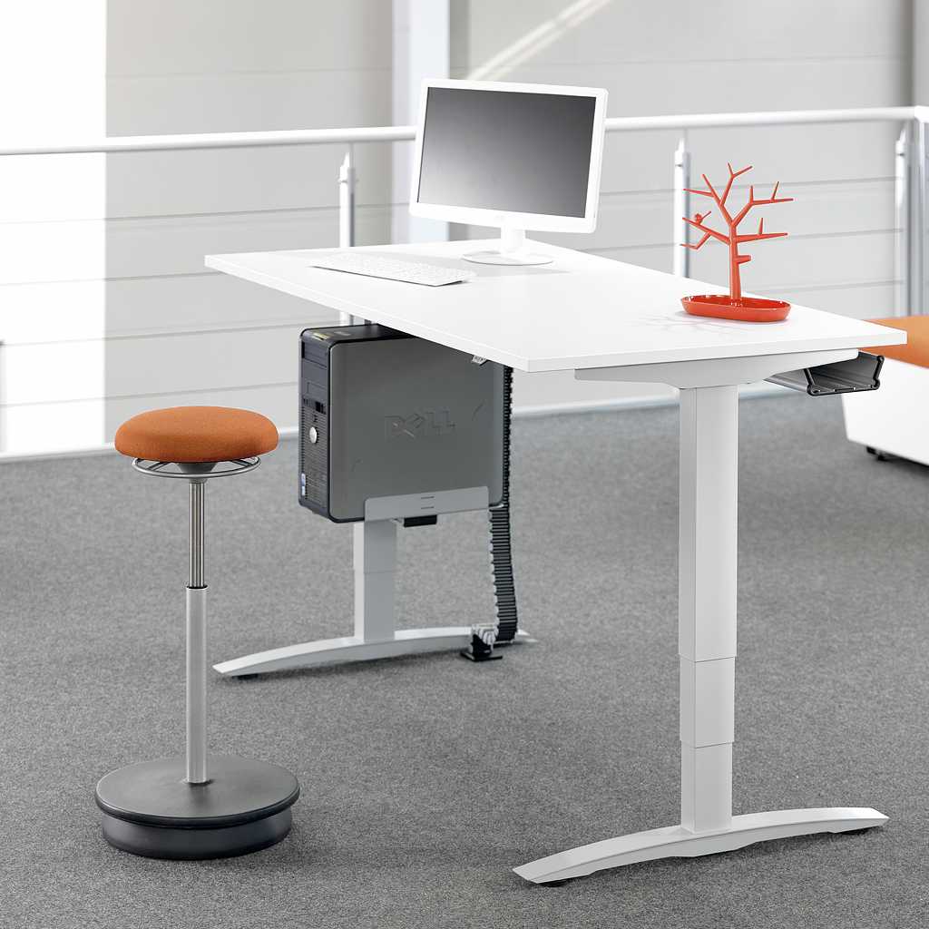 Stehhilfe im Büro Stehstuhl mit schwarzem Tellerfuß und orangenem Sitz vor einem weißen Stehschreibtisch im Büro. Auf dem Schreibtisch steht ein Monitor und orangene Deko. Grauer Teppichboden. Im Hintergrund ein Geländer.