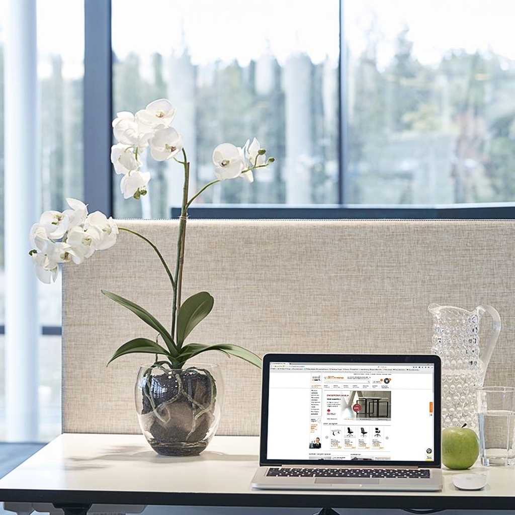Weiße Orchidee mit silbernem Topf. Kunstpflanze steht auf einem Schreibtisch neben einem Laptop. Dahinter eine graue Tischtrennwand.
