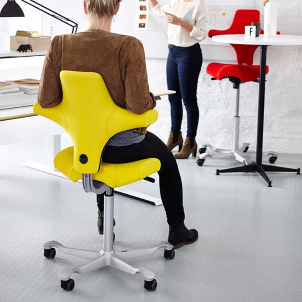 moderner Bürostuhl Person sitzt auf modernem Bürostuhl in gelber Farbe mit silbernerm Gestell und Fuß mit dem rücken zum Betrachter. Person ist am Schreibtisch und ist einer Person im Hintergrund zugewandt die neben einem roten Bürodrehstuhl steht.