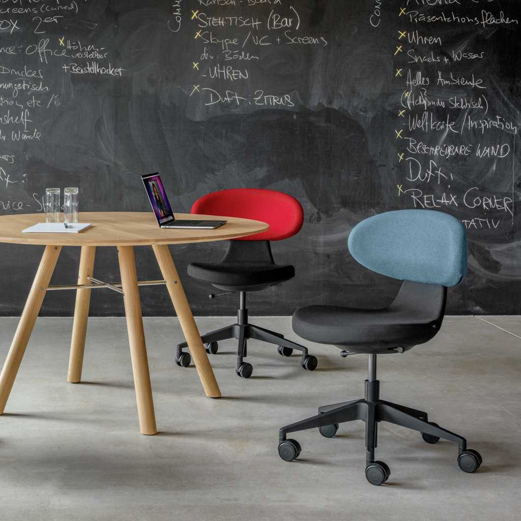 Besprechungsstuhl Zierliche Besprechungsstühle. Im Vordergrund ein Besprechungsstuhl mit schwarzem Sitzposter und kurzer mittelblauer Lehne auf schwarzem Drehkreuz mit Rollen. Im Hintergrund ein ähnlicher Stuhl, mit roter Lehne. Links seitlich dazwischen ein größerer Beistelltisch aus Holz. Im Hintergrund eine dunkelgraue Ziegelmauer. Betonboden.