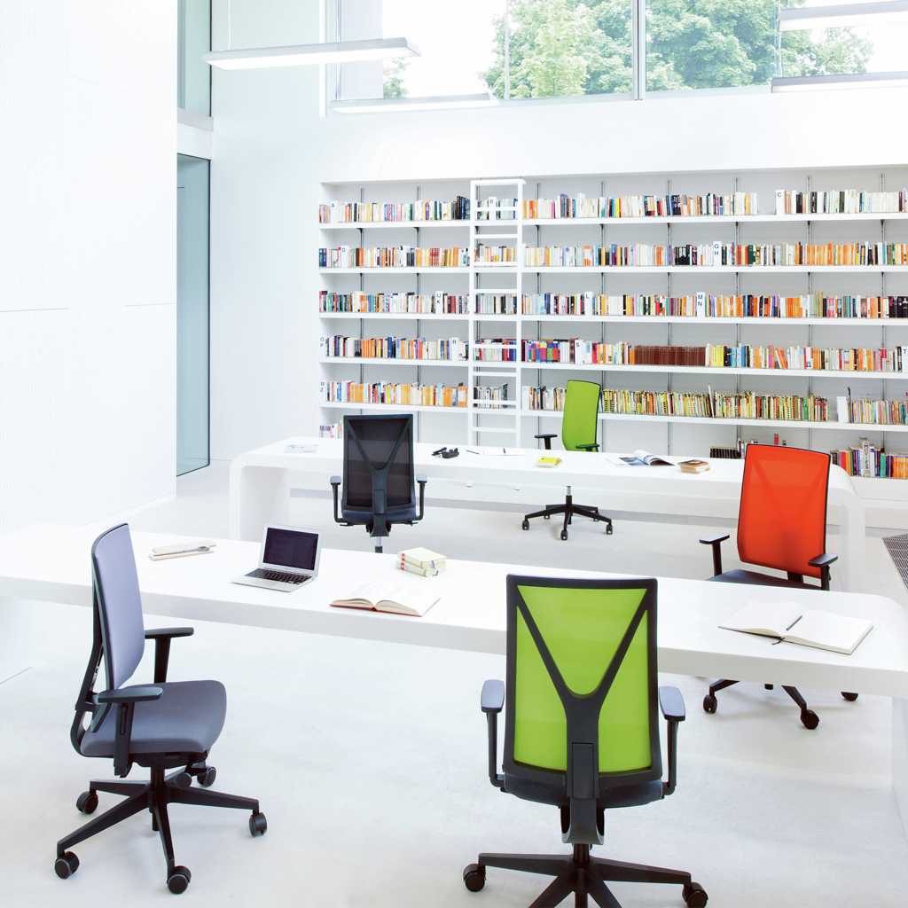 moderne Bürodrehstühle Um einen langen, weißen Tisch stehen mit Abstand vier Bürodrehstühle in den farben grau, grün, rot und schwarz. An einem weiteren Tisch steht noch eine einzelner grüner Drehstuhl. Im Hintergrund ist ein große Regalwand mit Büchern und einer Leiter. Oben eine große Glasfront.