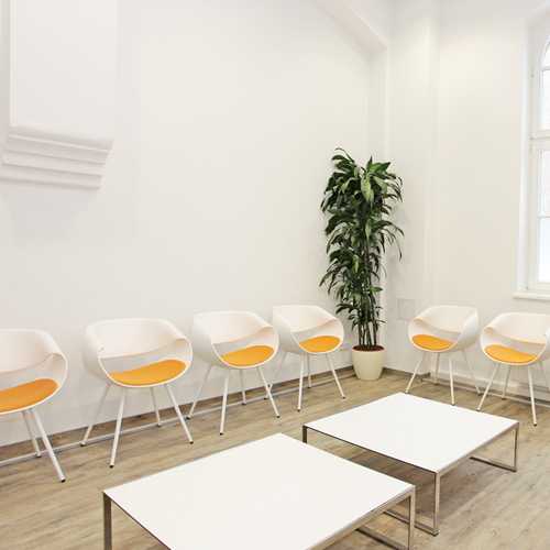 Sech weiße Design Wartestühle mit orangenem Polster. Im Vordergrund zwei flache, weiße Beistelltische. Im Hintergrund, in der Raumecke eine große Topfpflanze. 