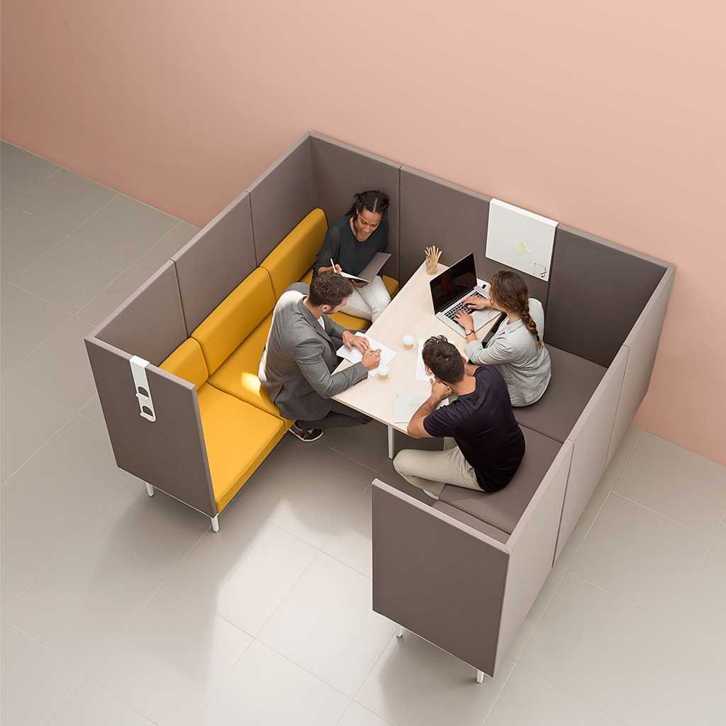 Blick von oben auf eine Sitzgruppe aus zwei Sofas. Links gelborange, rechts taupe; umrandet von einem taupefarbigen Sicht- und Akustikschutz. Um den Tisch in der Mitte sitzen vier Personen, eine mit Laptop.