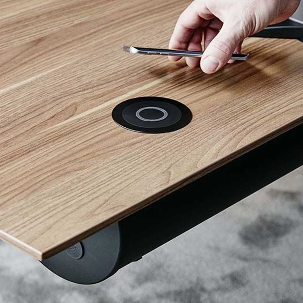 Induktionsladegerät für den Arbeitsplatz. EIne Ladestation in einen Holz-Schreibtisch integriert. Jemand legt das Handy auf der Induktionsstation ab. Eine Ladestation in einen Holz-Schreibtisch integriert. Jemand legt das Handy auf der Induktionsstation ab.