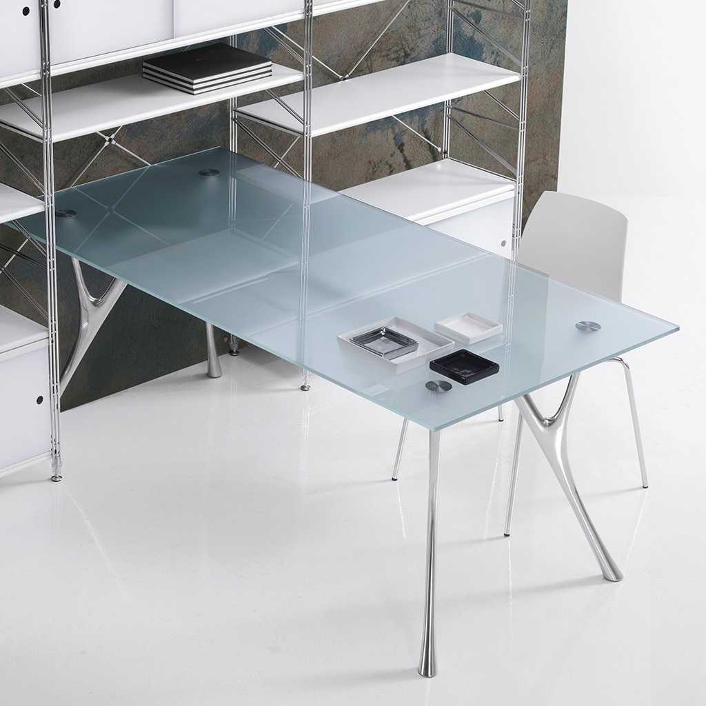 moderner Glas Schreibtisch moderner Schreibtisch mit Glasplatte auf silbernem Gestell. Auf der hinteren Seite grenz der Schreibtisch an ein filigranes Büroregal mit weißen Böden und silbernen Pfosten. Hinter dem Schreibtisch steht ein hellgrauer Bürostuhl mit Sitzschale.