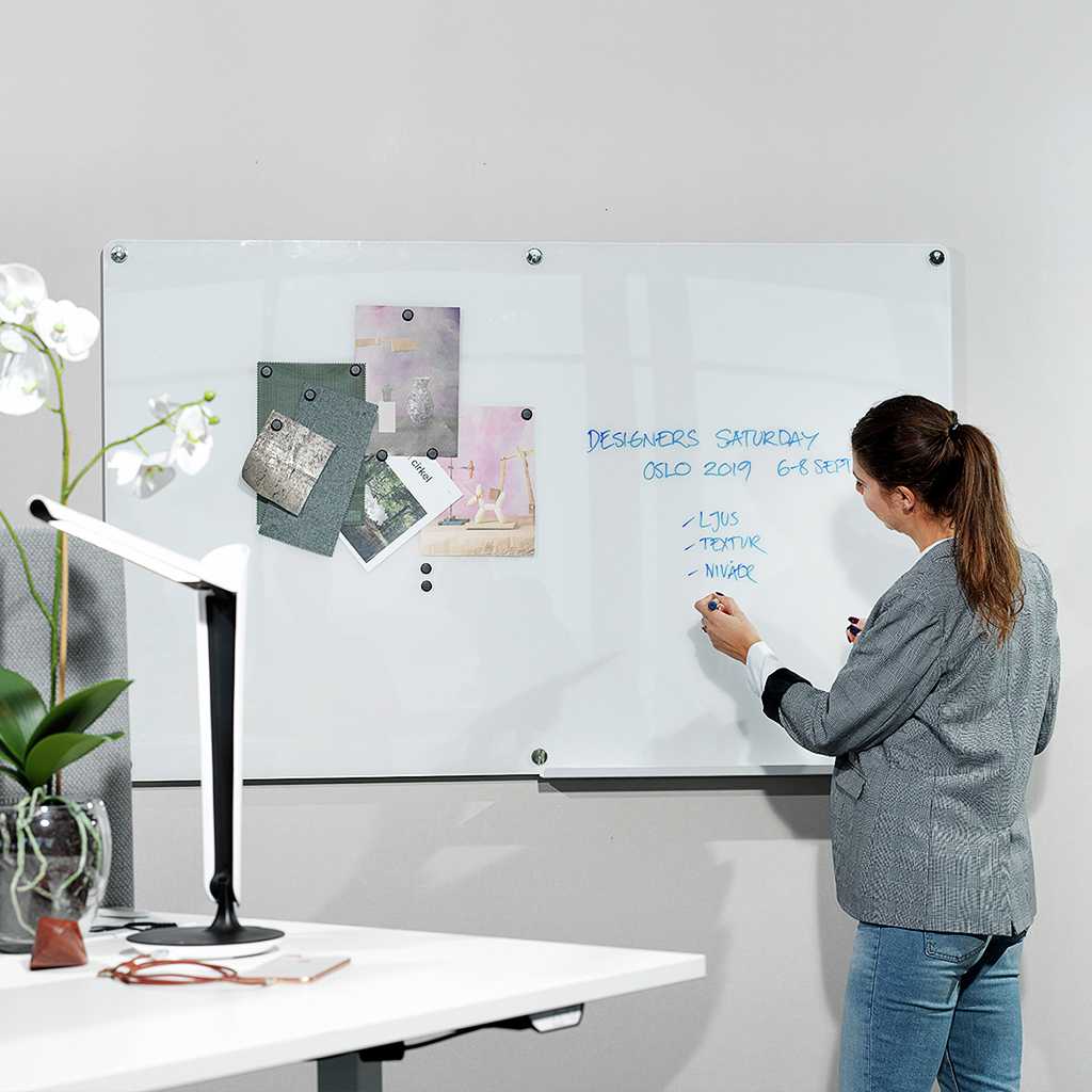 Whiteboard mit magnetischer Oberfläche an die Wand montiert. Auf dem Whiteboard Notizen und Bilder. Vor dem Whiteboard eine Frau die schreibt. Vor dem Whiteboard ein Arbeitsplatz mit einer Tischleuchte und einer Pflanze.