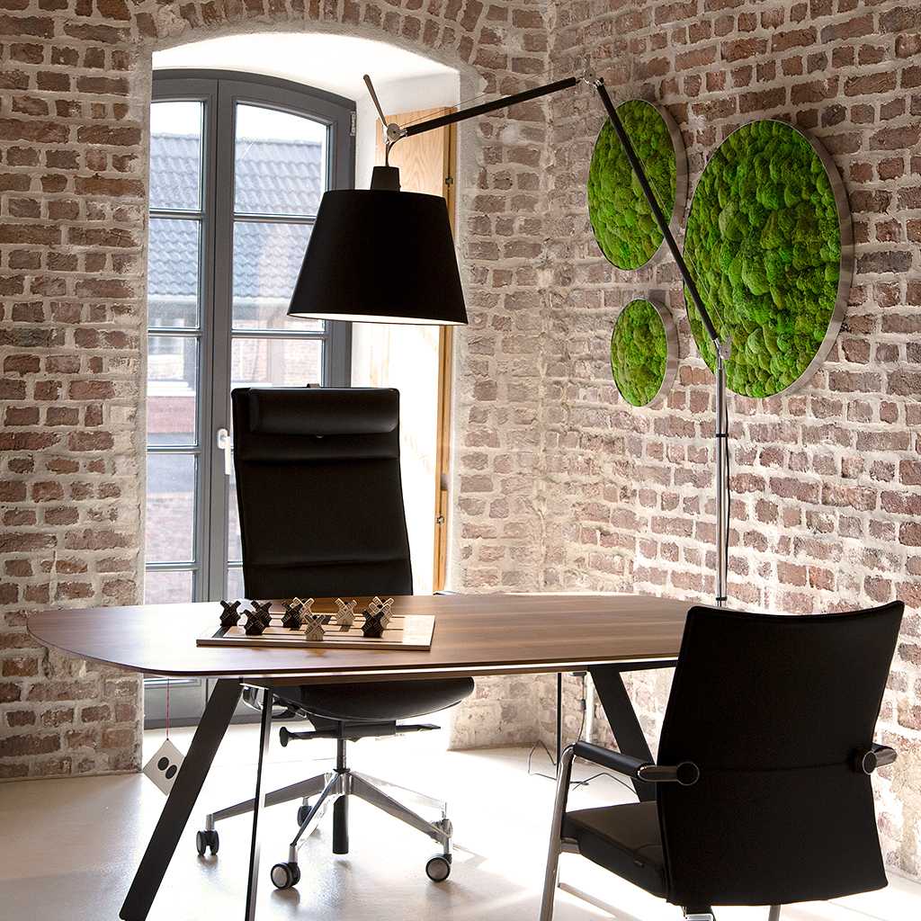 Design Stehlampe Große Design Stehleuchte mit schwarzem, runden Schirm. Gelenke aus poliertem Aluminium. Beleuchtet einen Schreibtisch aus dunklem Holz mit zwei Bürostühlen. An der Ziegelwand Moos-Akustikbilder.
