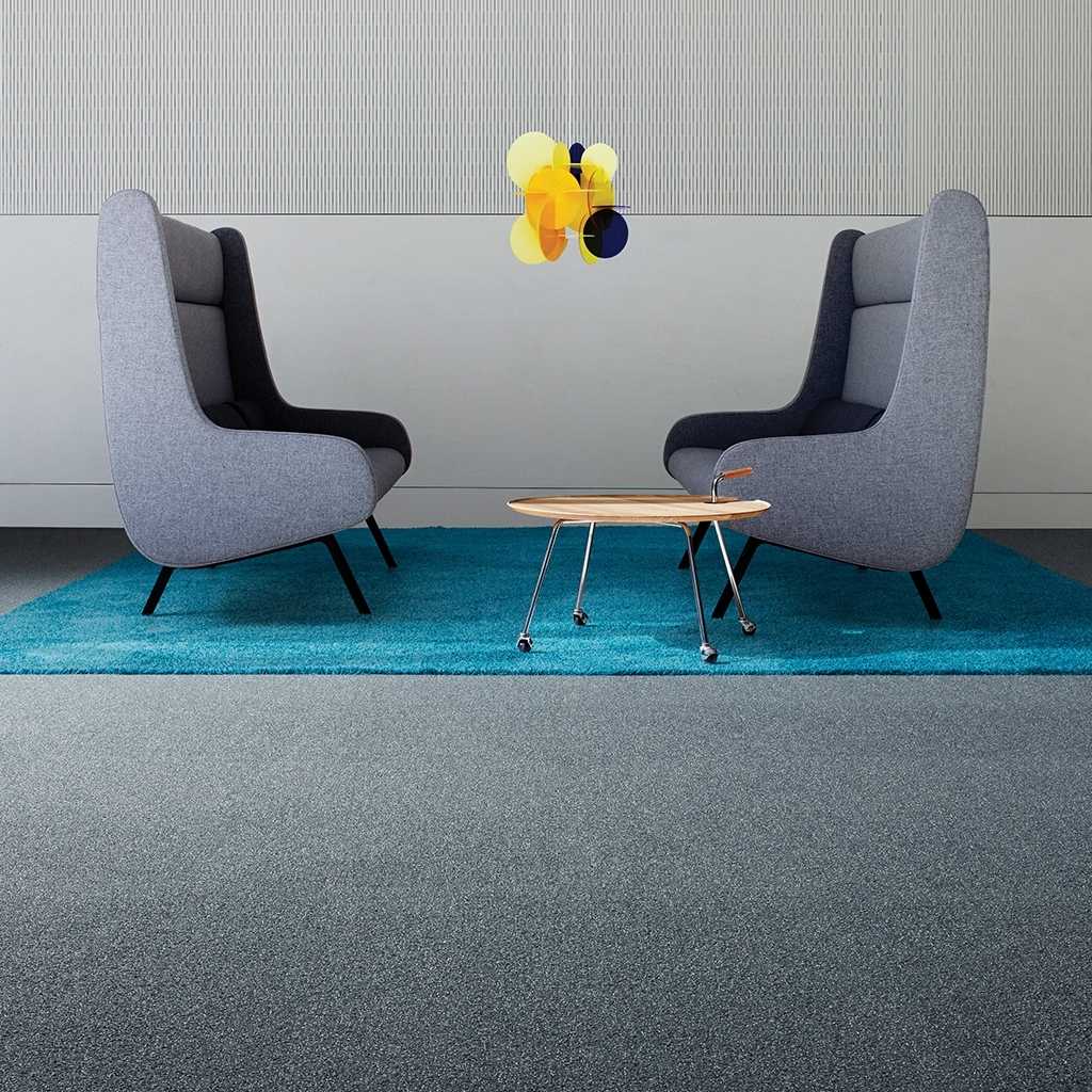 Besonders strapazierfähiger Teppichboden für Arbeitsräume in Blau. Auf dem Teppich stehen zwei große, graue Loungesessel gegenüber. Ein kleiner Beistelltisch steht neben den Sesseln. Eine bunte Pendelleuchte hängt zwischen den Sesseln.