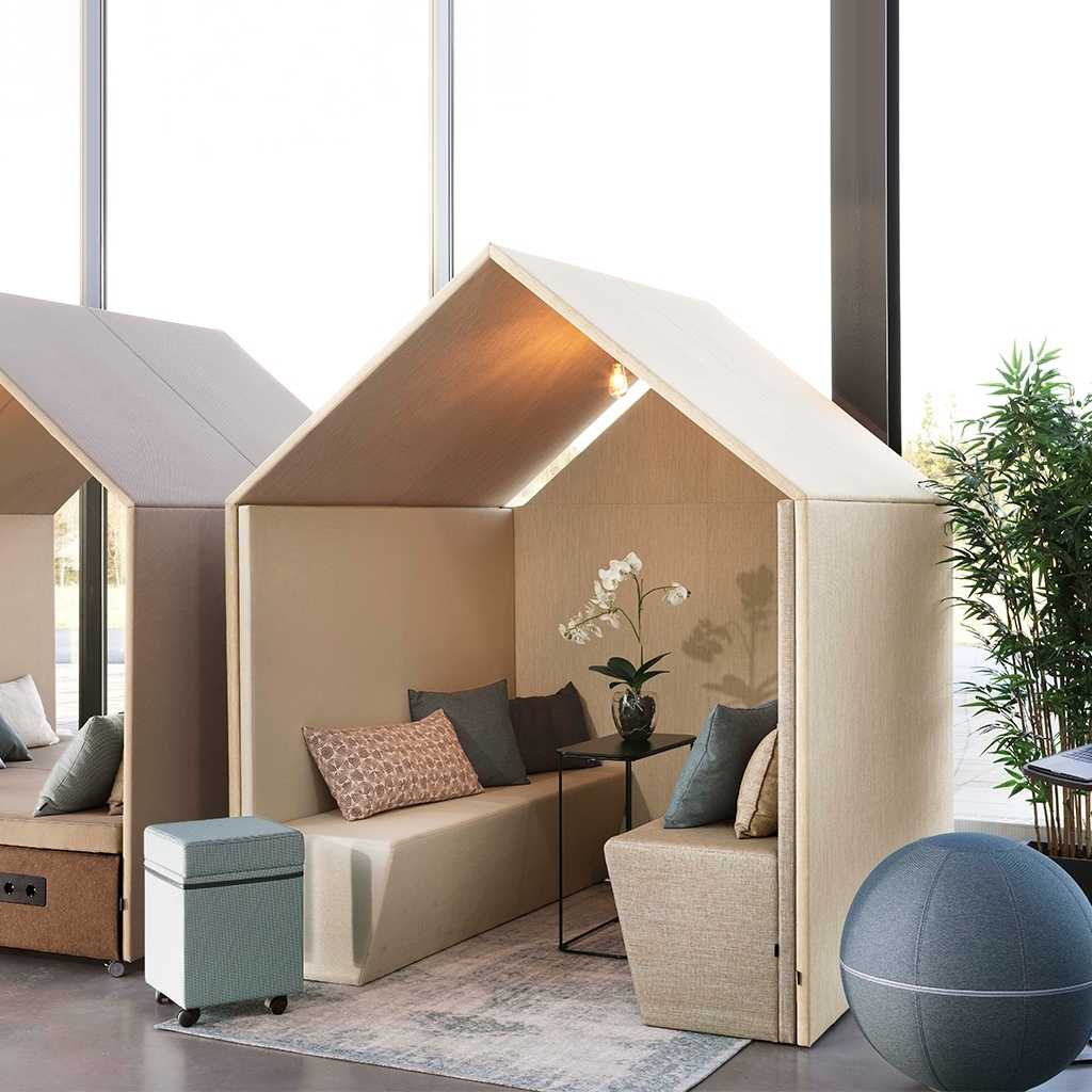 Meetingpoint in Hüttenform Kleine beige Hüttenform. Innen zwei Bänke und ein schmaler schwarzer Tisch. Im Hintergrund Pflanzen .Vorne rechts ein Sitzball, links ein kleiner Rollhocker.