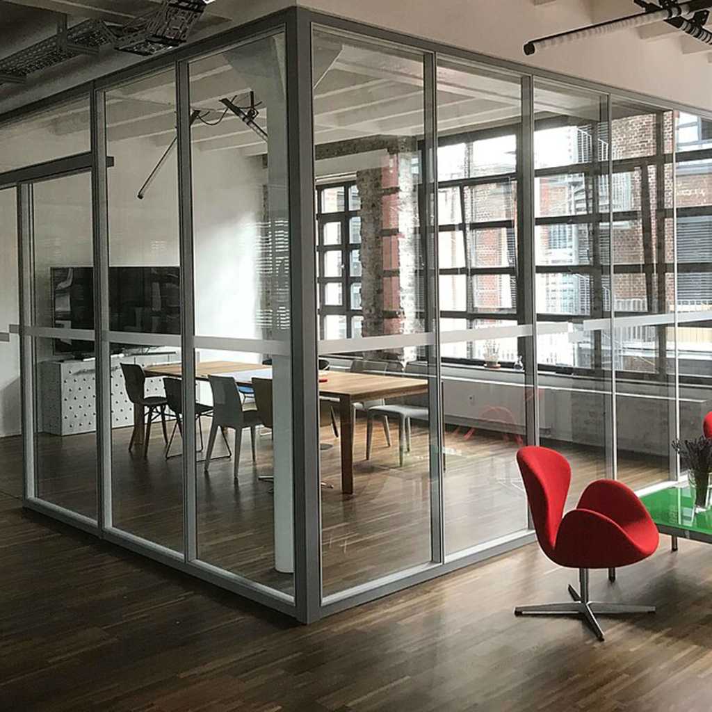 Trennwandsystem aus Glas Glastrennwand als Raummtrennung über Eck. Hinter der Glaswand ein großer Konferenztisch mit Stühlen und ein großer Bildschirm.