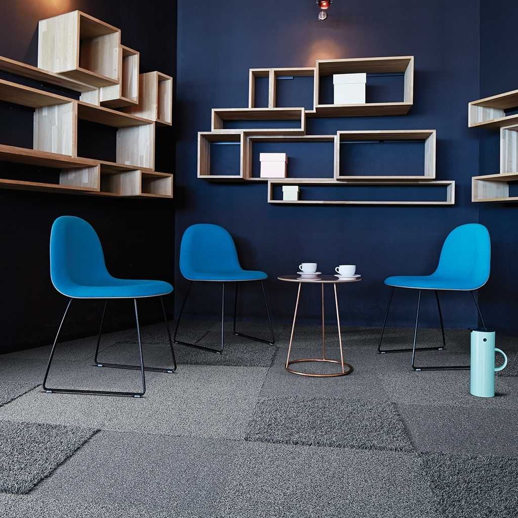 Teppichfliesen in unterschiedlichen grau Tönen. Im Raum stehen drei blaue Besucherstühle und ein kleiner Beistelltisch. An der Wand hänegn Design Holzregale. 