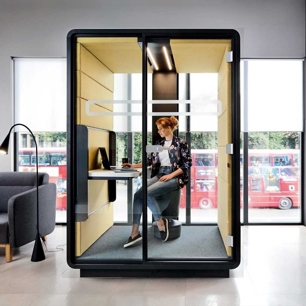 Arbeitsraum im Raum Blick in einen in sich geschlossenen Arbeitsraum in einem größeren Raum. Im Raum ein Arbeitsplatz mit Schreibtisch und Stuhl, daran eine Person. Der Boden im Raumsystem ist grau. Zwei der Wände sind gelb, ebenso die Decke. Man blickt durch eine Glastrennwand mit Tür in den Raum hinein und durch eine weitere Glastrennwand hinten wieder hinnaus.