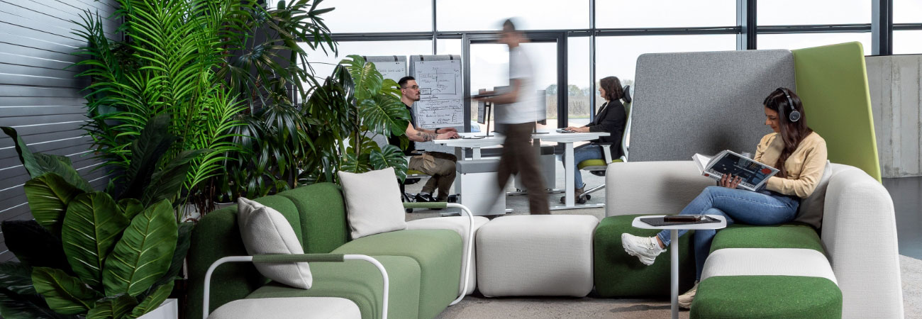 Bürolounge mit Pflanzen und modularen Sofas; zwei Personen arbeiten, vorne liest eine Frau mit Kopfhörern.